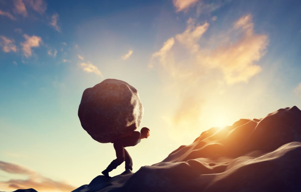 Man carrying a boulder on his back while walking up a mountain.